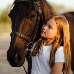 Paulina Rasińska Fotografia - Portret dziewczynki w białej sukience, obejmującej czarnego konia z uprzężą. Delikatne światło podkreśla bliskość i więź między dzieckiem a zwierzęciem.