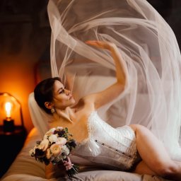 Paulina Rasińska Fotografia - Kobieta w białym gorsecie i welonie leży na łóżku, trzymając bukiet kwiatów. W tle lampka nocna i ciemna ściana. Ujęcie z sesji w stylu boudoir.