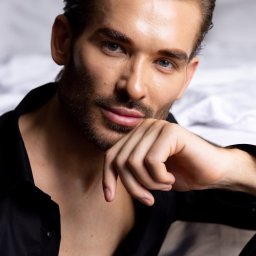 Paulina Rasińska Fotografia - Portret mężczyzny z lekkim zarostem, w rozpiętej czarnej koszuli, opierającego brodę na dłoni. Gładko zaczesane włosy, jasne oczy, miękkie światło. Białe tło.