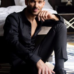 Paulina Rasińska Fotografia - Mężczyzna w czarnym stroju siedzi na podłodze w pokoju hotelowym, opierając brodę na dłoni. W tle widoczne łóżko i lampka nocna. Ujęcie z dołu.