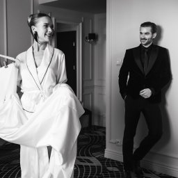 Paulina Rasińska Fotografia - Elegancka sesja w hotelu: kobieta w szlafroku trzyma suknię, mężczyzna w smokingu opiera się o ścianę. Czarno-biała fotografia z wyrafinowanym klimatem.