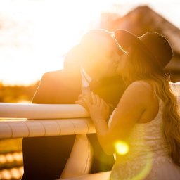 Paulina Rasińska Fotografia - Zakochana para w kapeluszach całuje się przy białym płocie w promieniach zachodzącego słońca. Romantyczna sesja w plenerze z budynkiem w tle.