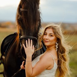 Paulina Rasińska Fotografia - Uśmiechnięta blondynka w białej sukni pozuje z czarnym koniem na tle łąki. Delikatne światło podkreśla detale sukni i biżuterii. Wietrzna pogoda.