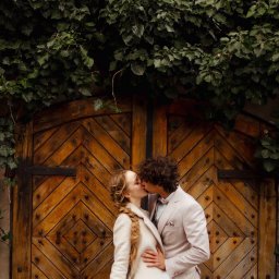 Paulina Rasińska Fotografia - Zakochana para w pastelowych strojach całuje się na tle starych, drewnianych wrót obrośniętych bluszczem. Romantyczna sesja w plenerze, ciepłe światło.