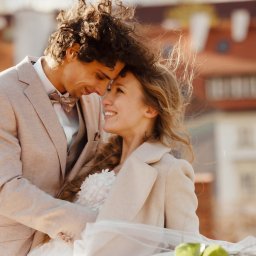 Paulina Rasińska Fotografia - Uśmiechnięta para młoda w beżowych strojach, przytulona na tle miejskiej zabudowy z czerwonymi dachami. Delikatne światło podkreśla romantyczny nastrój.