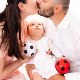 Paulina Rasińska Fotografia - Rodzice całujący się nad niemowlęciem w białym ubranku, które trzyma dwie małe piłki: biało-czarną i czerwono-czarną. Dziecko patrzy w górę. Matka w różowej sukience, ojciec w białej koszuli.