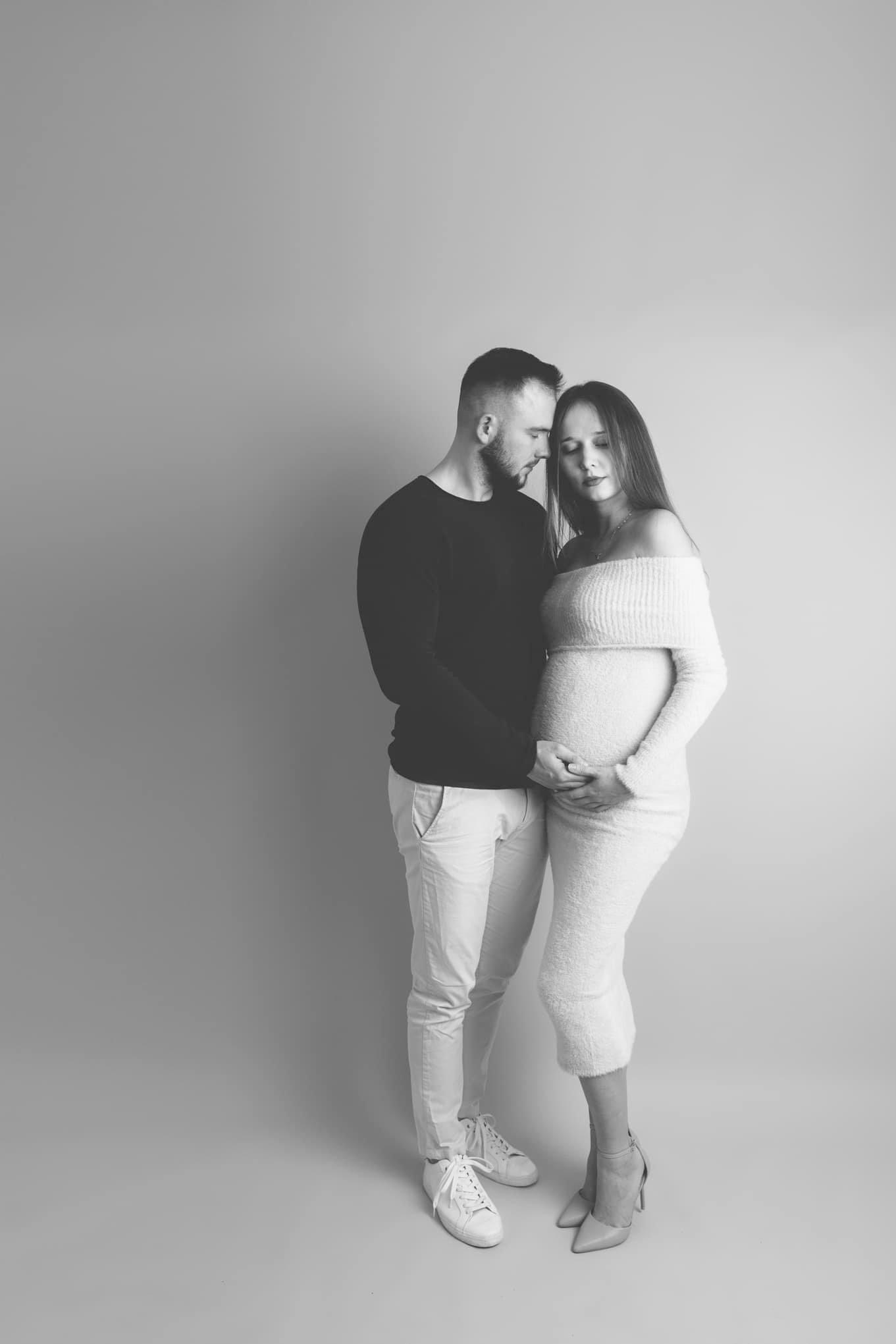 Czarno-białe zdjęcie pary: mężczyzna w czarnym swetrze i jasnych spodniach trzyma ręce na brzuchu kobiety w białej, obcisłej sukience z odkrytymi ramionami i butach na obcasie, oboje z zamkniętymi...