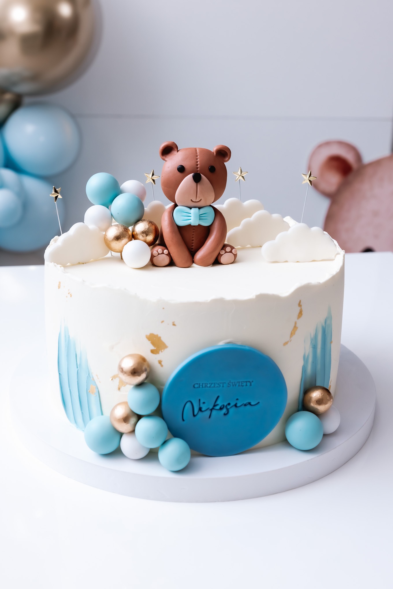 Tort na chrzest z białym kremem, niebieskimi akcentami i figurką brązowego misia w niebieskiej muszce, otoczony błękitnymi, białymi i złotymi kulkami oraz dekoracjami w kształcie chmurek i gwiazdek.