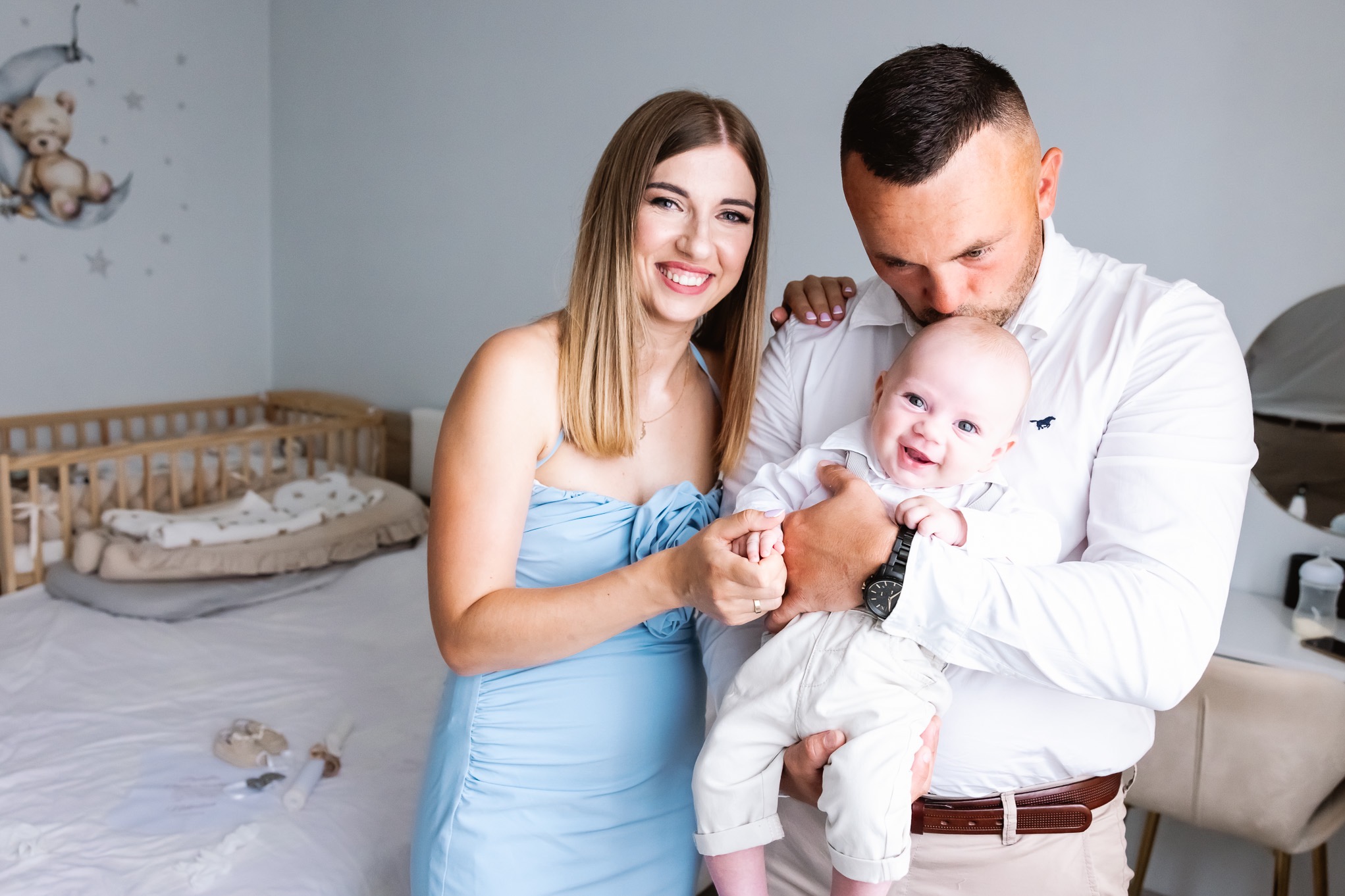 Uśmiechnięta rodzina: mama w błękitnej sukience trzyma za rękę tatę całującego w głowę uśmiechniętego niemowlaka w białym ubranku na tle pokoju dziecięcego z łóżeczkiem i naklejką misia na księżycu.
