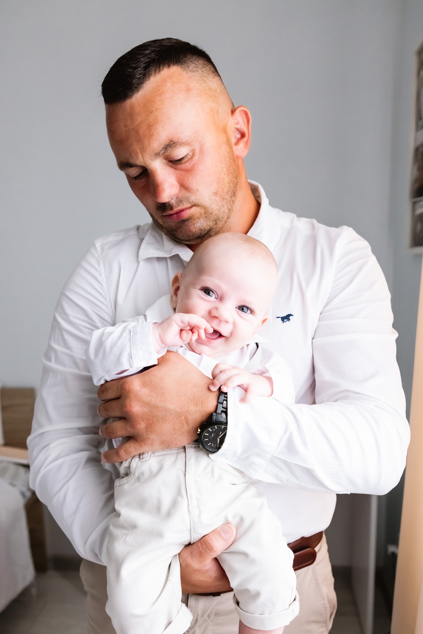 Uśmiechnięty niemowlak w jasnych ubraniach trzymany przez ojca w białej koszuli z wyhaftowanym logo konika.