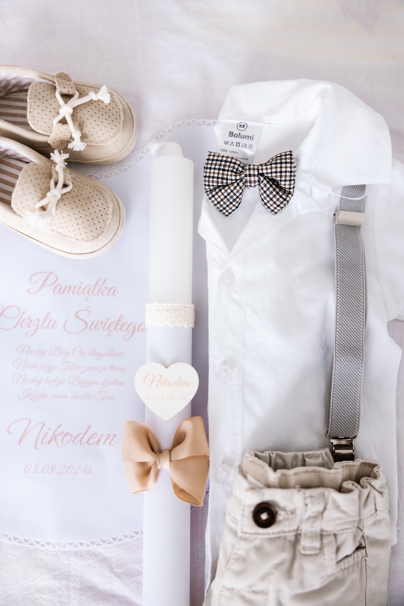 Kompozycja z pamiątką Chrztu Świętego w Busko-Zdroju: biała świeca z imieniem Nikodem i datą 03.08.2024, karta z napisem 'Pamiątka Chrztu Świętego', eleganckie buciki, biała koszula z muszką...