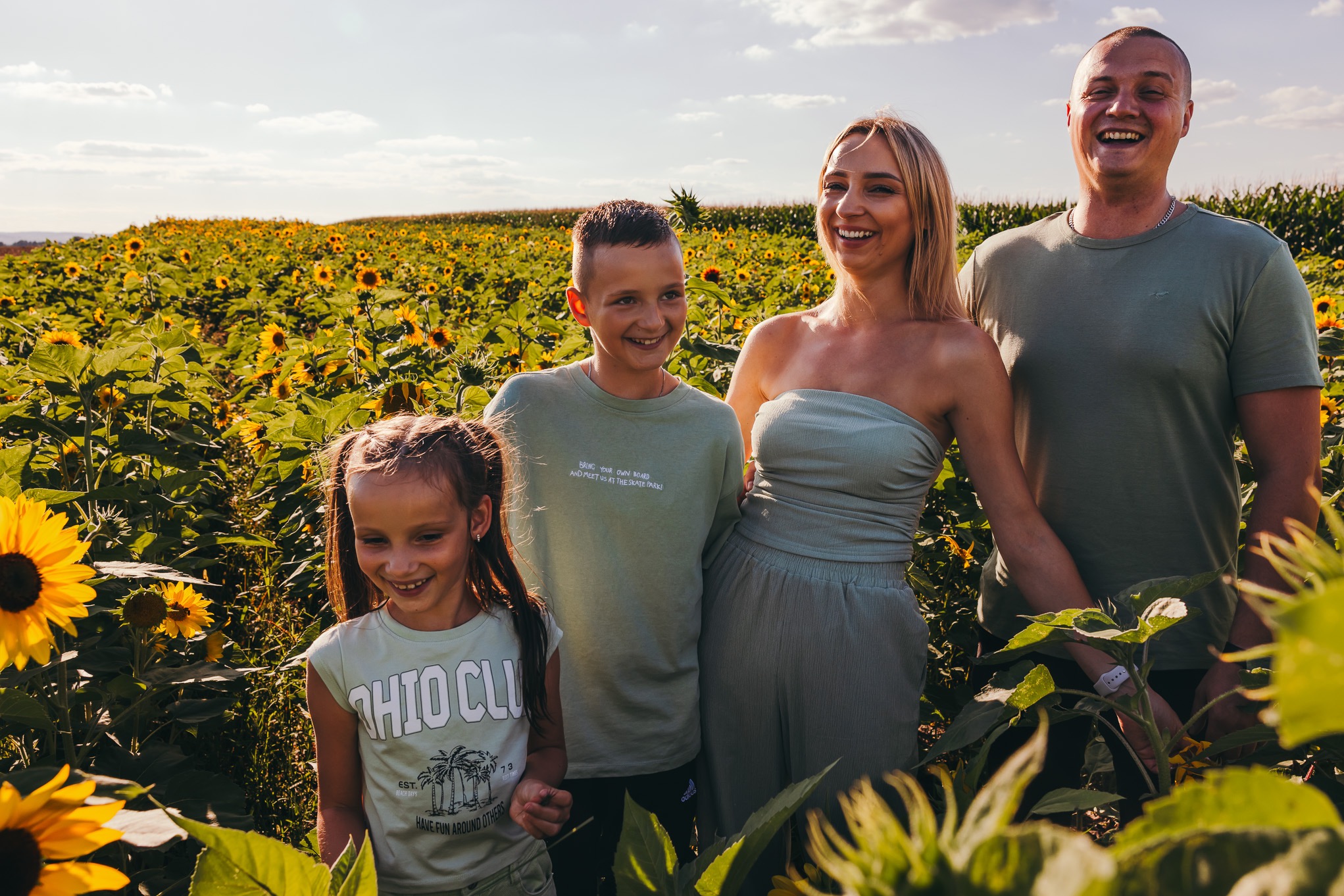 Uśmiechnięta rodzina (mama, tata, syn i córka) pozuje w polu słoneczników w słoneczny dzień. Rodzice i syn stoją, a córka lekko pochylona. W tle widoczne niebo i odległe drzewa.