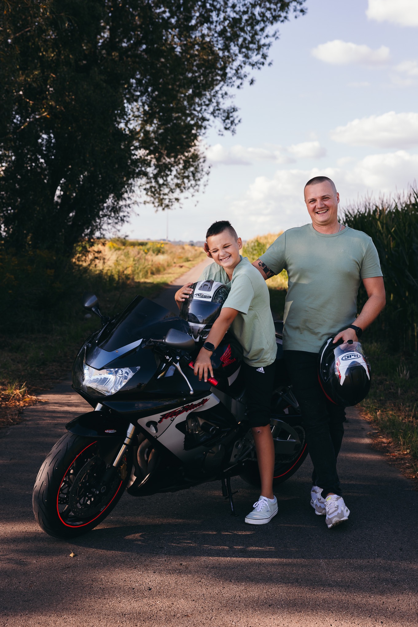Uśmiechnięty ojciec z synem pozują na tle motocykla sportowego na wiejskiej drodze, obaj trzymają kaski.