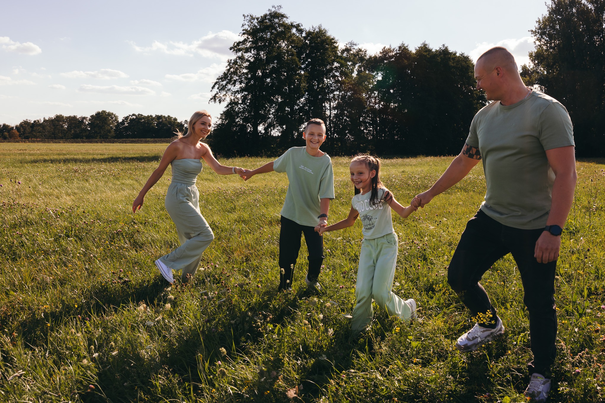 Rodzina trzymająca się za ręce, spacerująca po łące w słoneczny dzień. Mama w pistacjowym kombinezonie, tata w zielonej koszulce i czarnych spodniach, syn w zielonej koszulce, córka w jasnej...