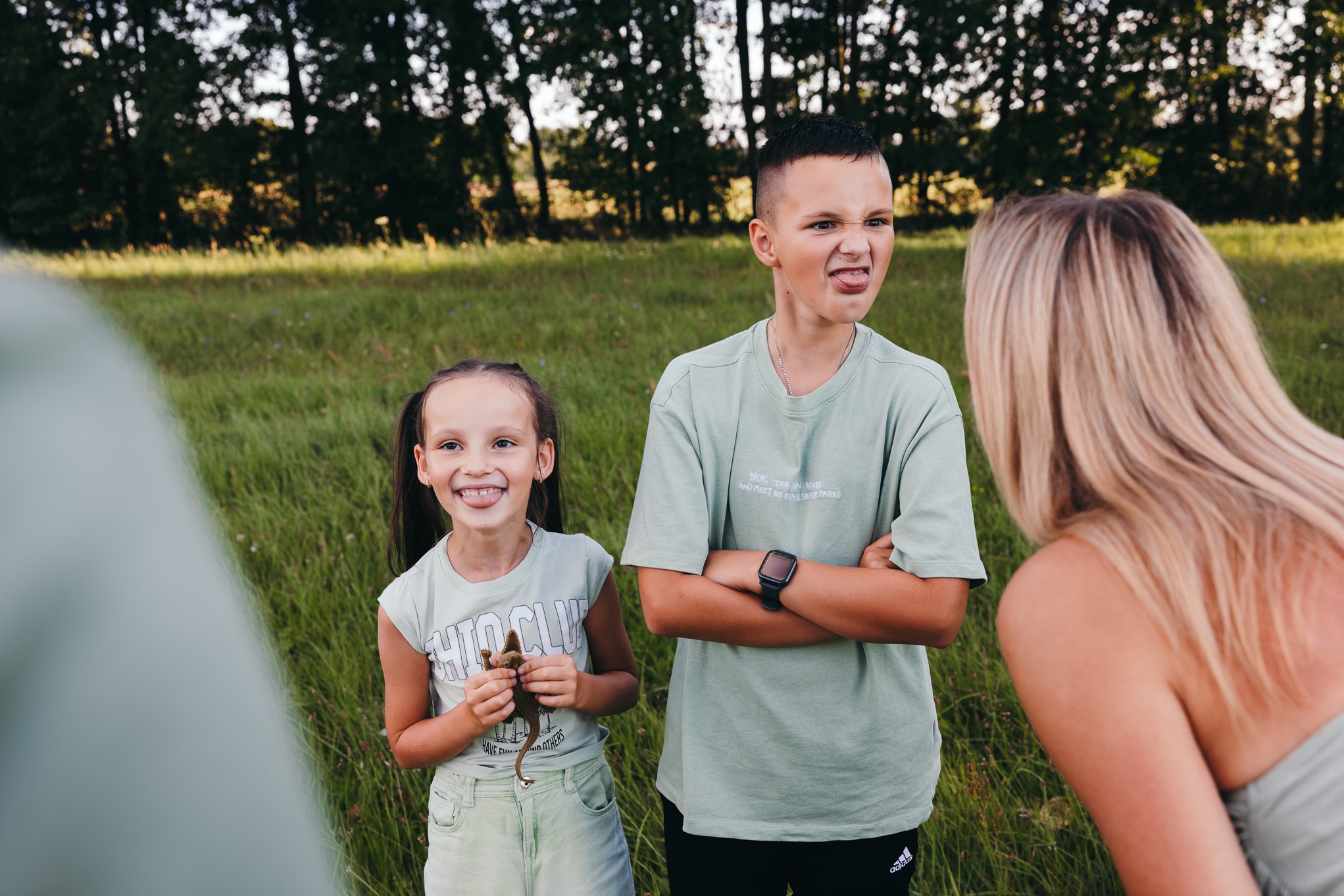 Uśmiechnięta dziewczynka trzymająca plastikowego dinozaura, obok chłopiec z wyciągniętym językiem i kobieta z blond włosami, na tle zielonej trawy i drzew.