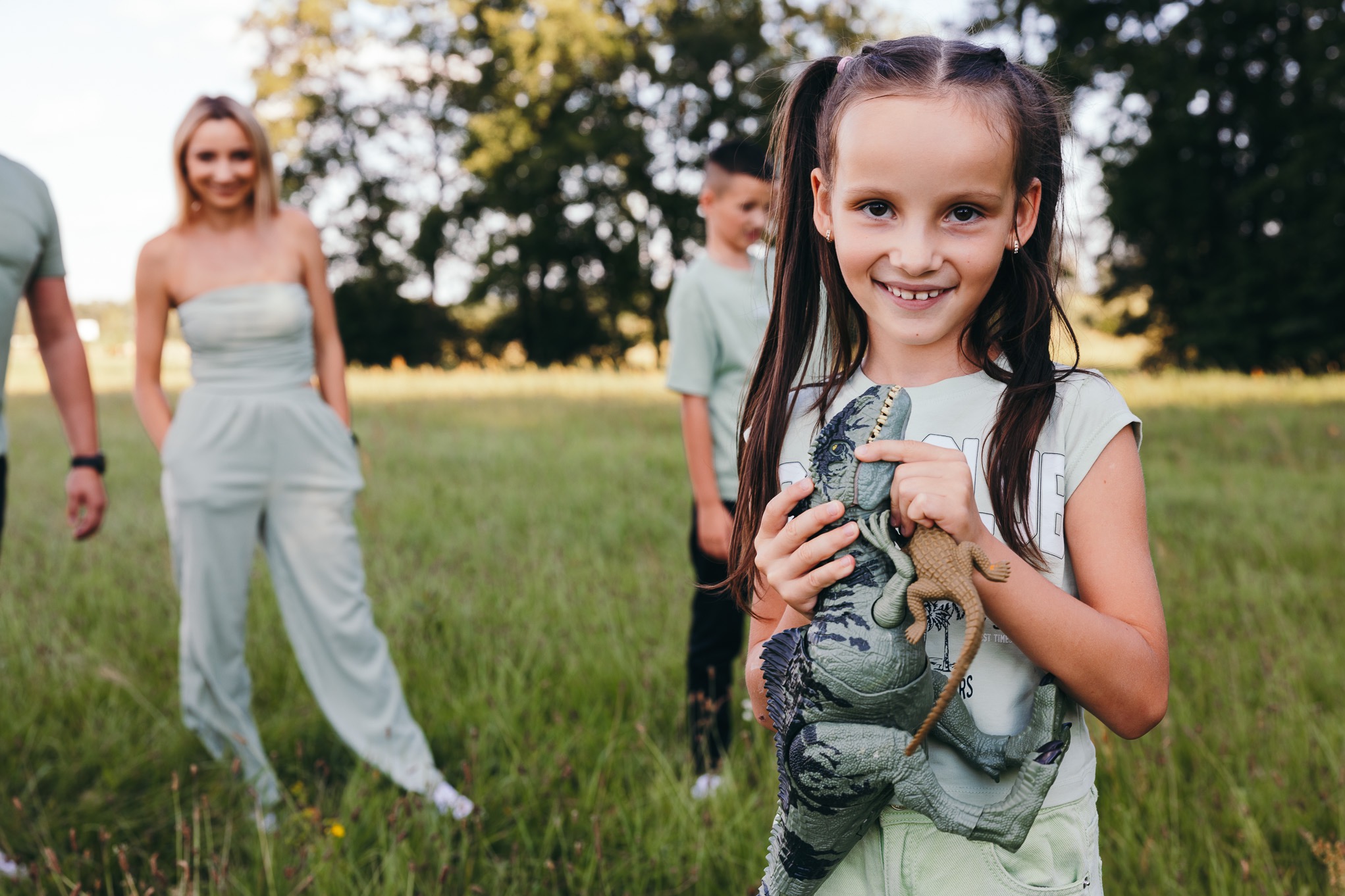 Uśmiechnięta dziewczynka z warkoczykami trzymająca dwa dinozaury na tle rodziny w pasujących pastelowych strojach, stojącej na łące.