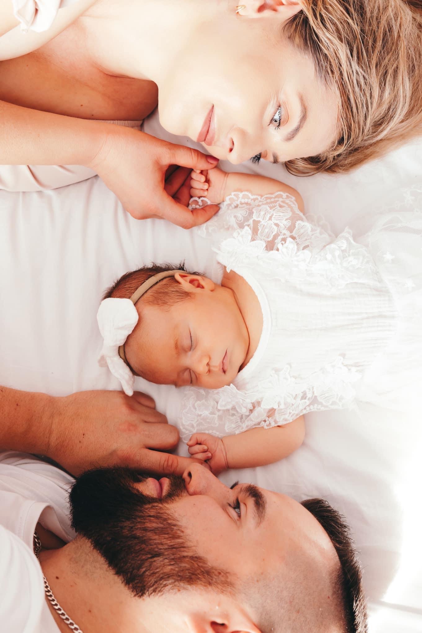 Noworodek w koronkowej sukience śpiący między rodzicami leżącymi na białym prześcieradle, delikatne oświetlenie, widoczna bransoletka na ręce matki i łańcuszek na szyi ojca.