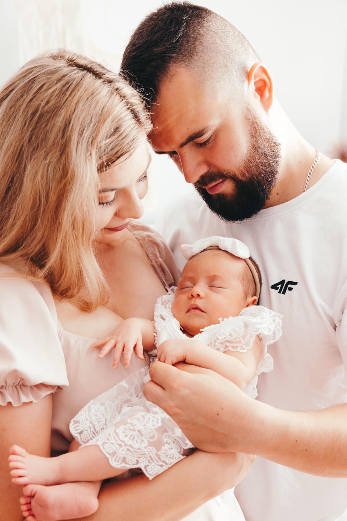 Młoda matka i ojciec czule spoglądają na śpiące niemowlę ubrane w białą koronkową sukienkę i opaskę z kokardką, ojciec ma na sobie białą koszulkę z logo 4F.