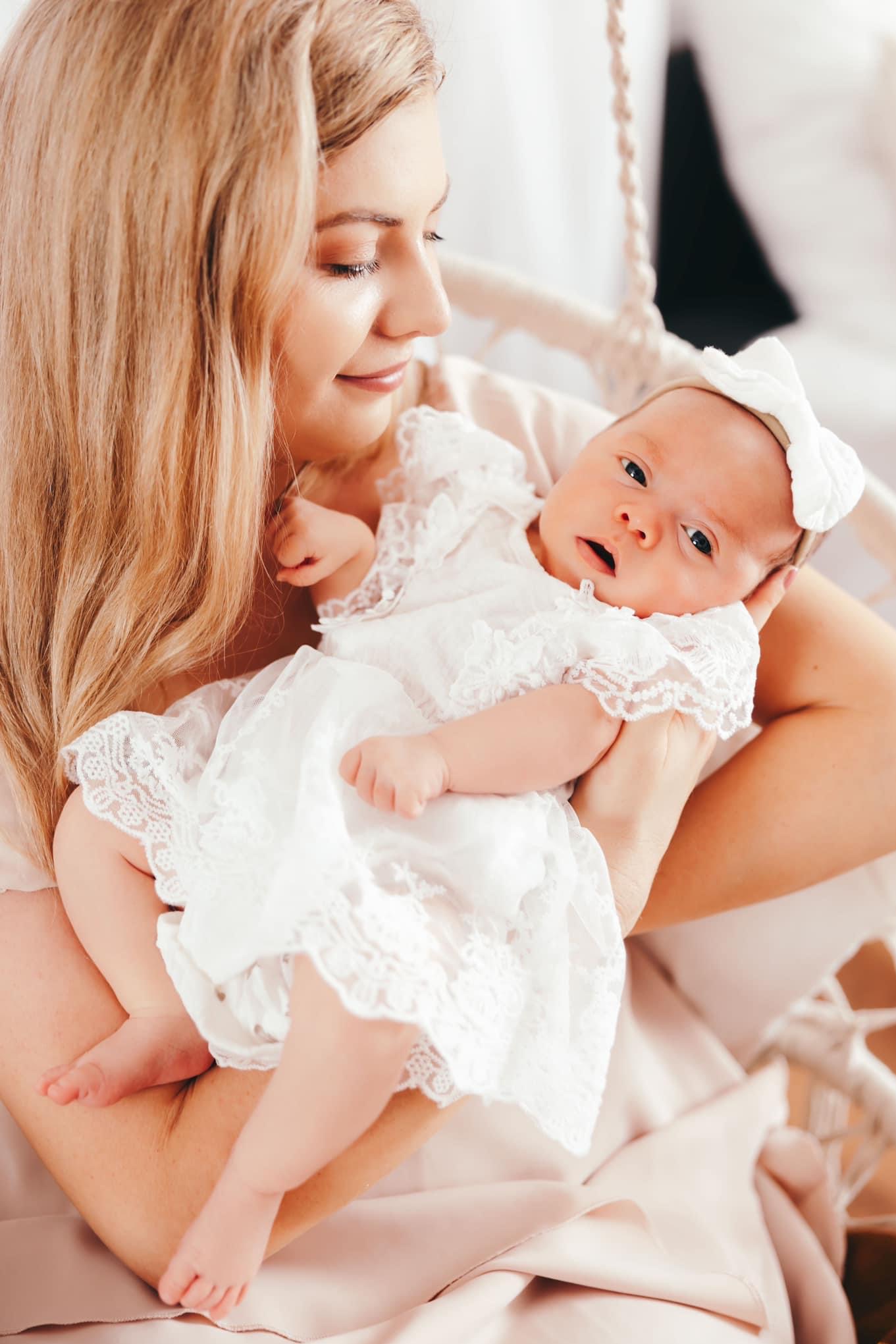 Blondwłosa matka trzyma na rękach niemowlę ubrane w białą, koronkową sukienkę i opaskę z kokardą, patrzące w obiektyw.