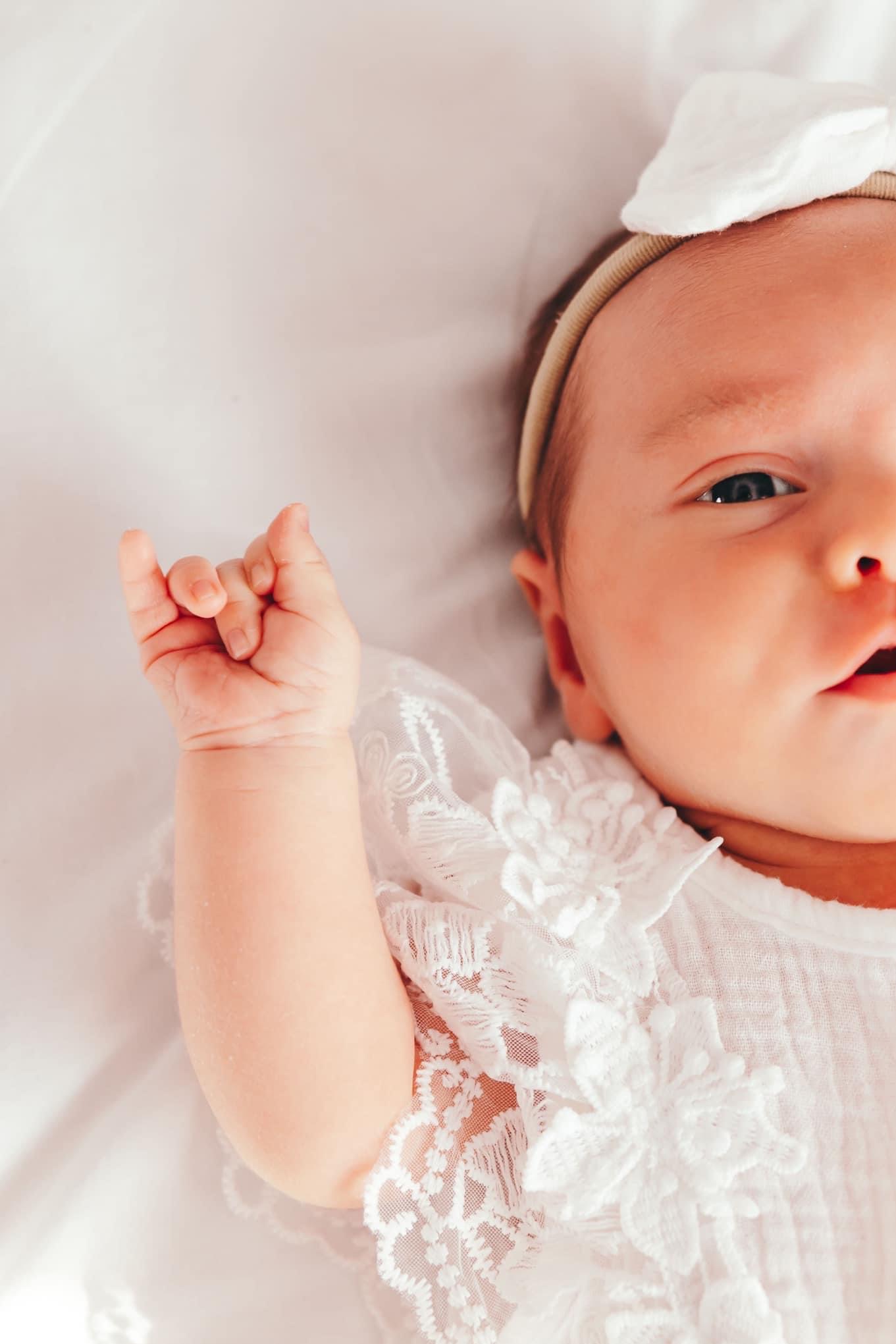 Portret noworodka w białej koronkowej sukience z kokardką na głowie, leżącego na białym tle, z uniesioną rączką.