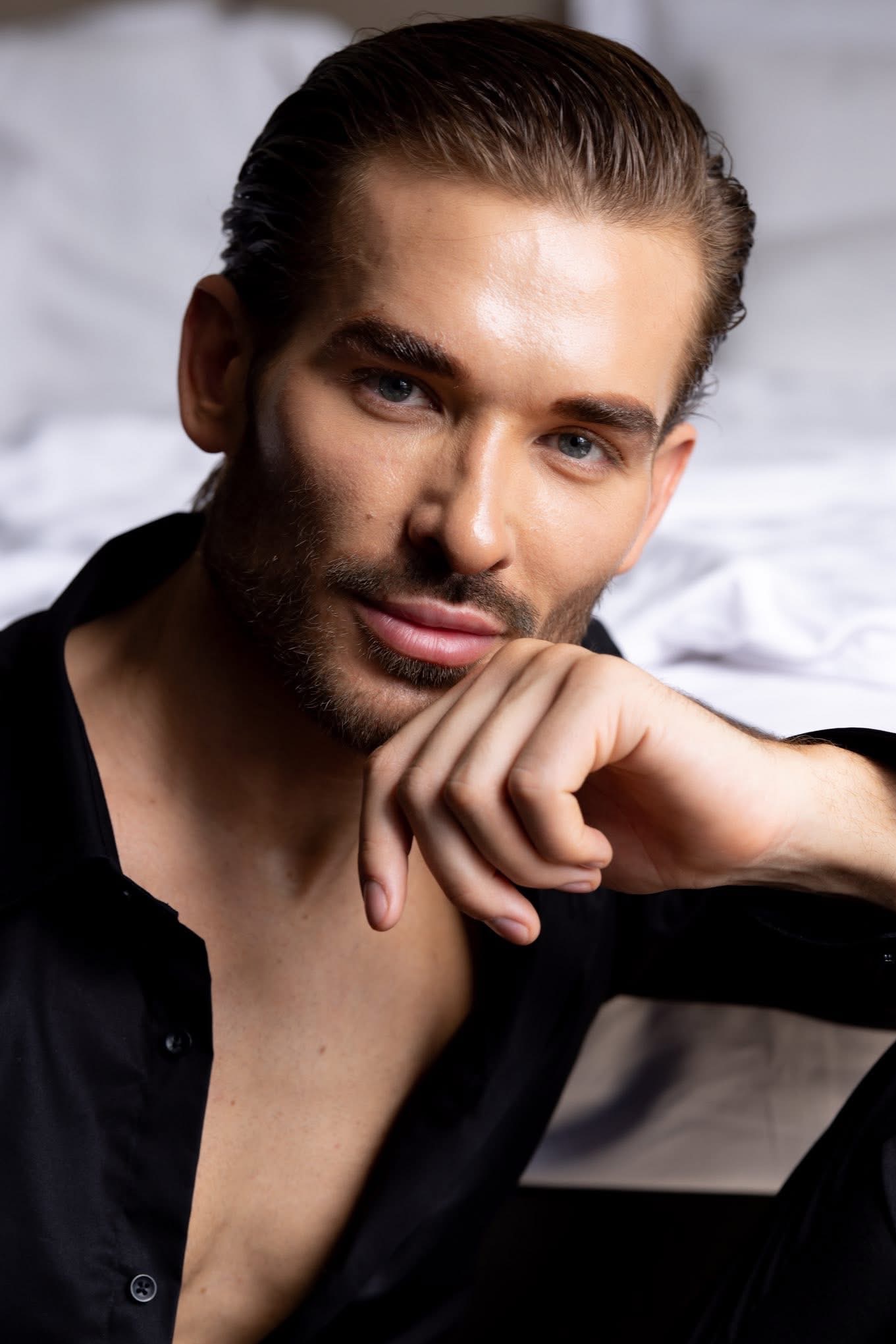 Portret mężczyzny z lekkim zarostem, w rozpiętej czarnej koszuli, opierającego brodę na dłoni. Gładko zaczesane włosy, jasne oczy, miękkie światło. Białe tło.