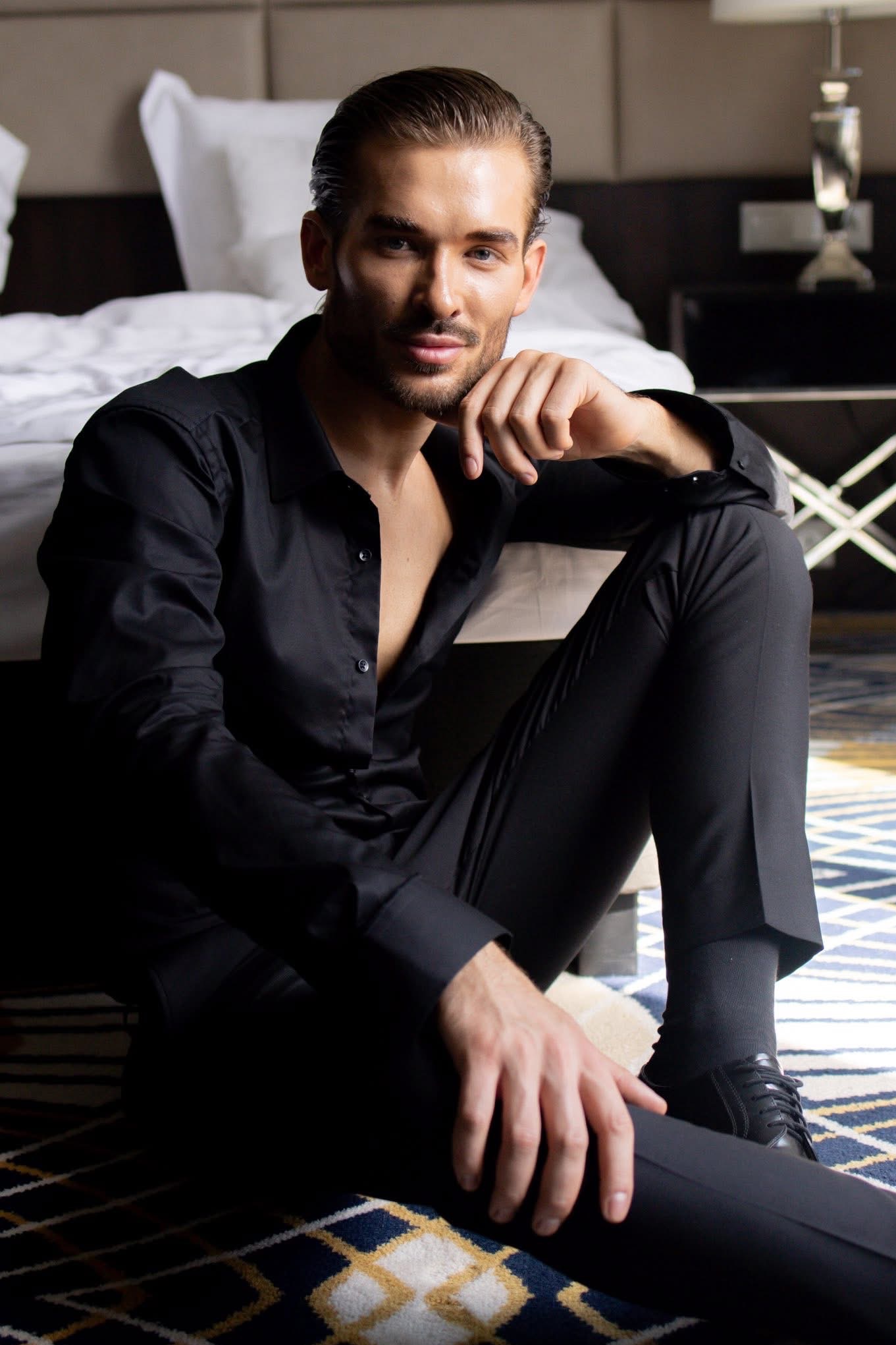 Mężczyzna w czarnym stroju siedzi na podłodze w pokoju hotelowym, opierając brodę na dłoni. W tle widoczne łóżko i lampka nocna. Ujęcie z dołu.