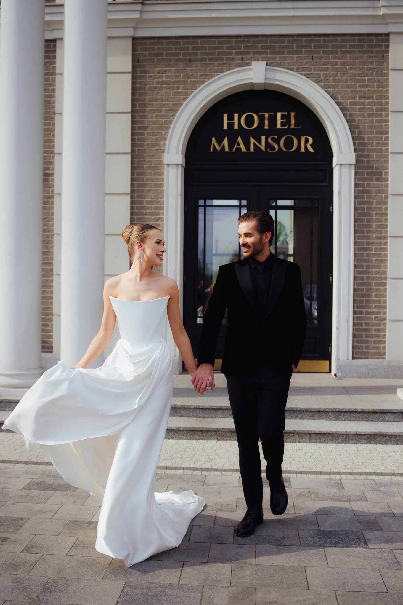 Elegancka para młoda, on w czarnym smokingu, ona w białej sukni, trzymają się za ręce na tle Hotel Mansor. Uśmiechnięci, w ruchu, w słoneczny dzień.