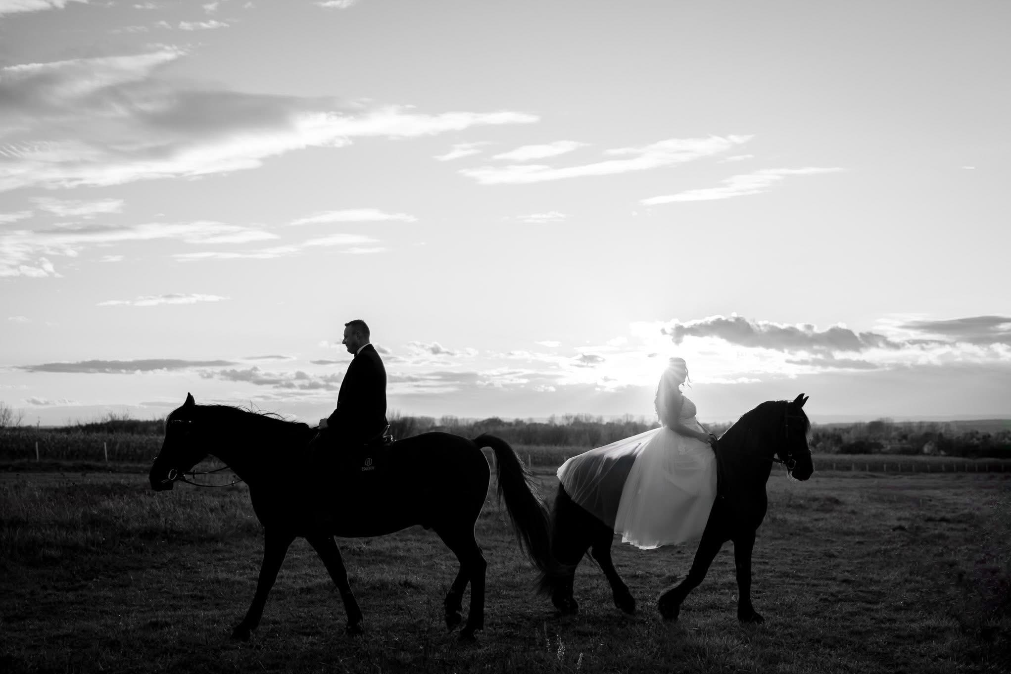 Czarno-białe zdjęcie pary młodej na koniach na tle zachodzącego słońca i pochmurnego nieba. Pan młody w garniturze, panna młoda w sukni ślubnej. Romantyczna sesja w plenerze.