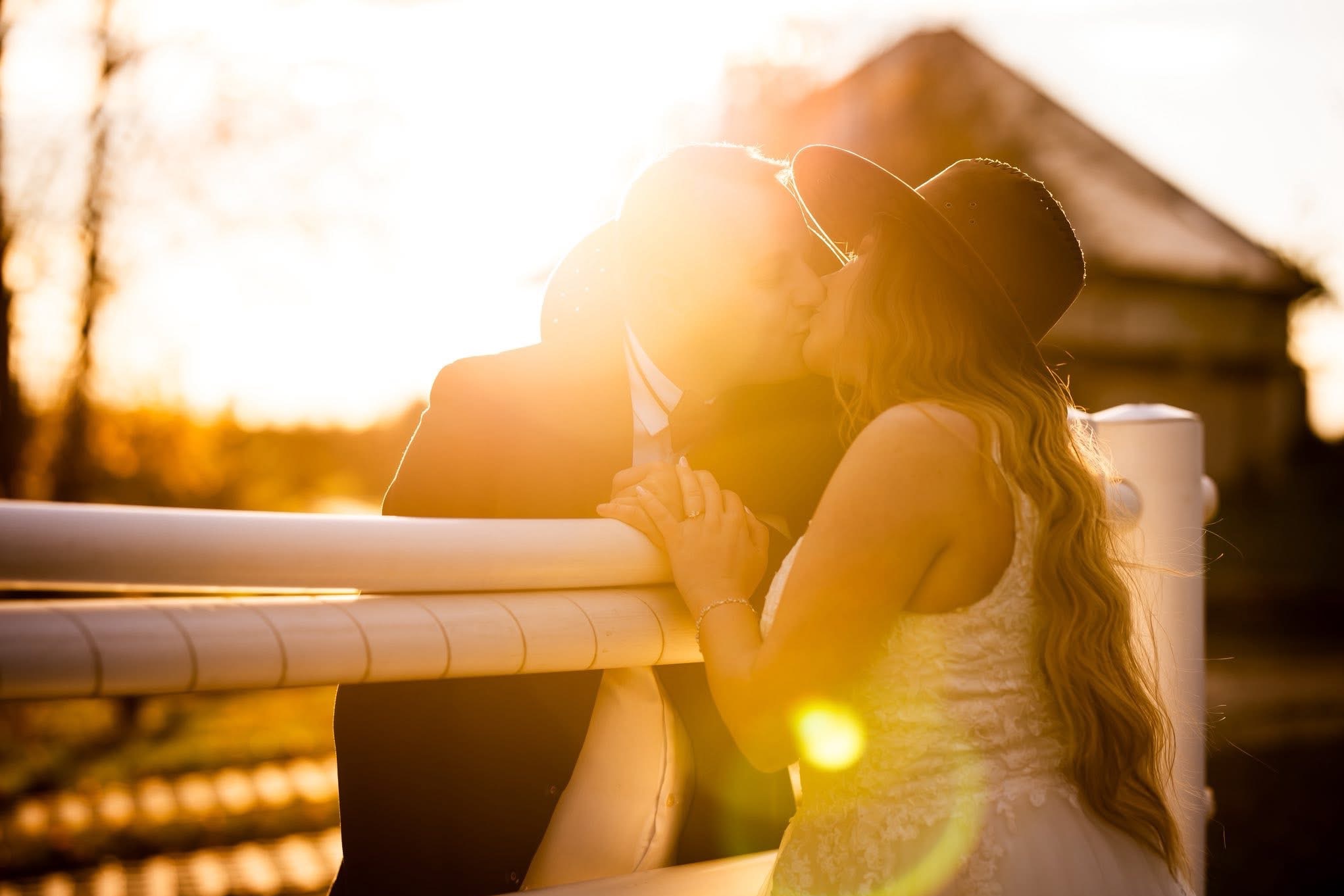 Zakochana para w kapeluszach całuje się przy białym płocie w promieniach zachodzącego słońca. Romantyczna sesja w plenerze z budynkiem w tle.