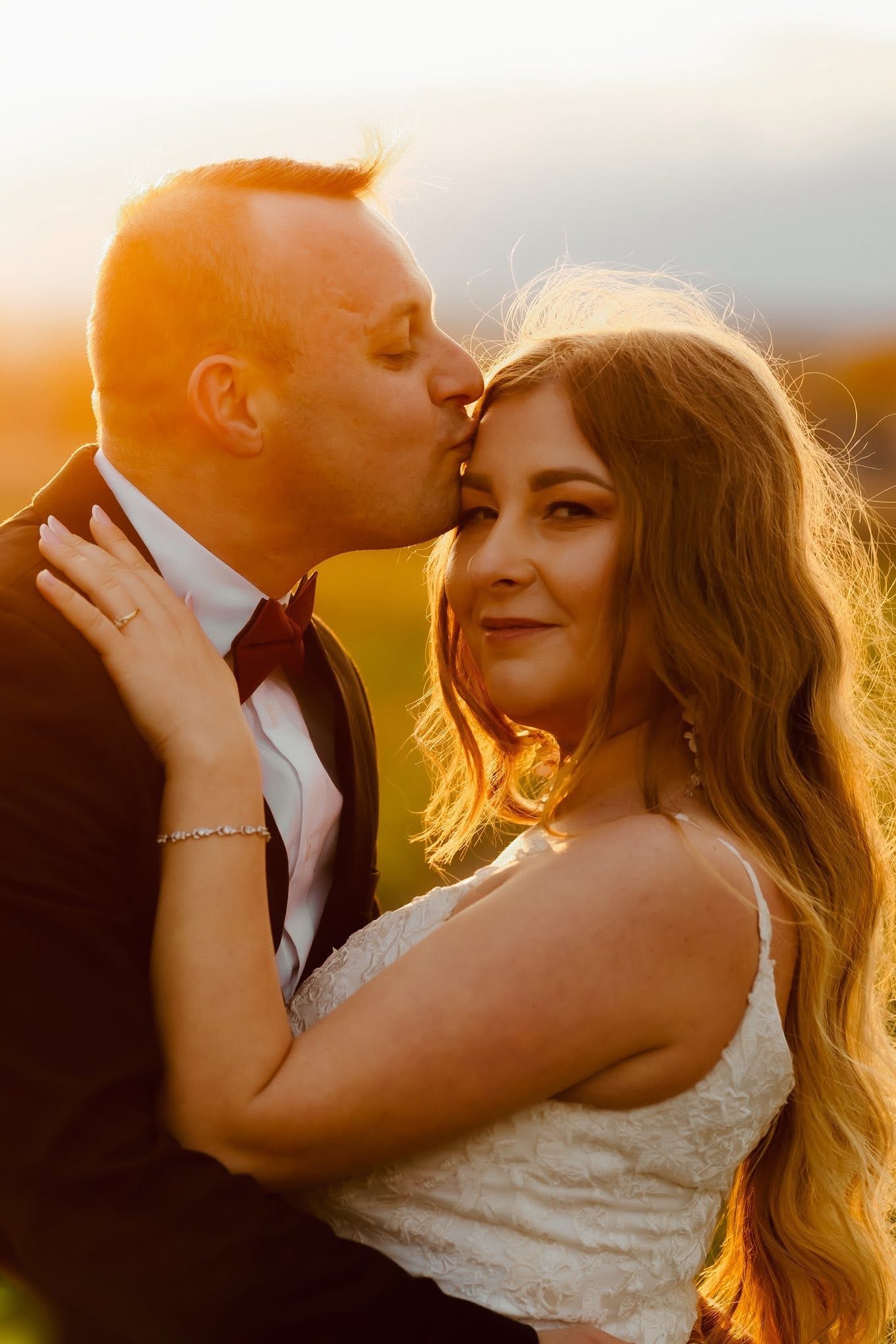 Uśmiechnięta panna młoda w objęciach pana młodego, który całuje ją w czoło, w ciepłym świetle zachodzącego słońca. Romantyczna sesja plenerowa.