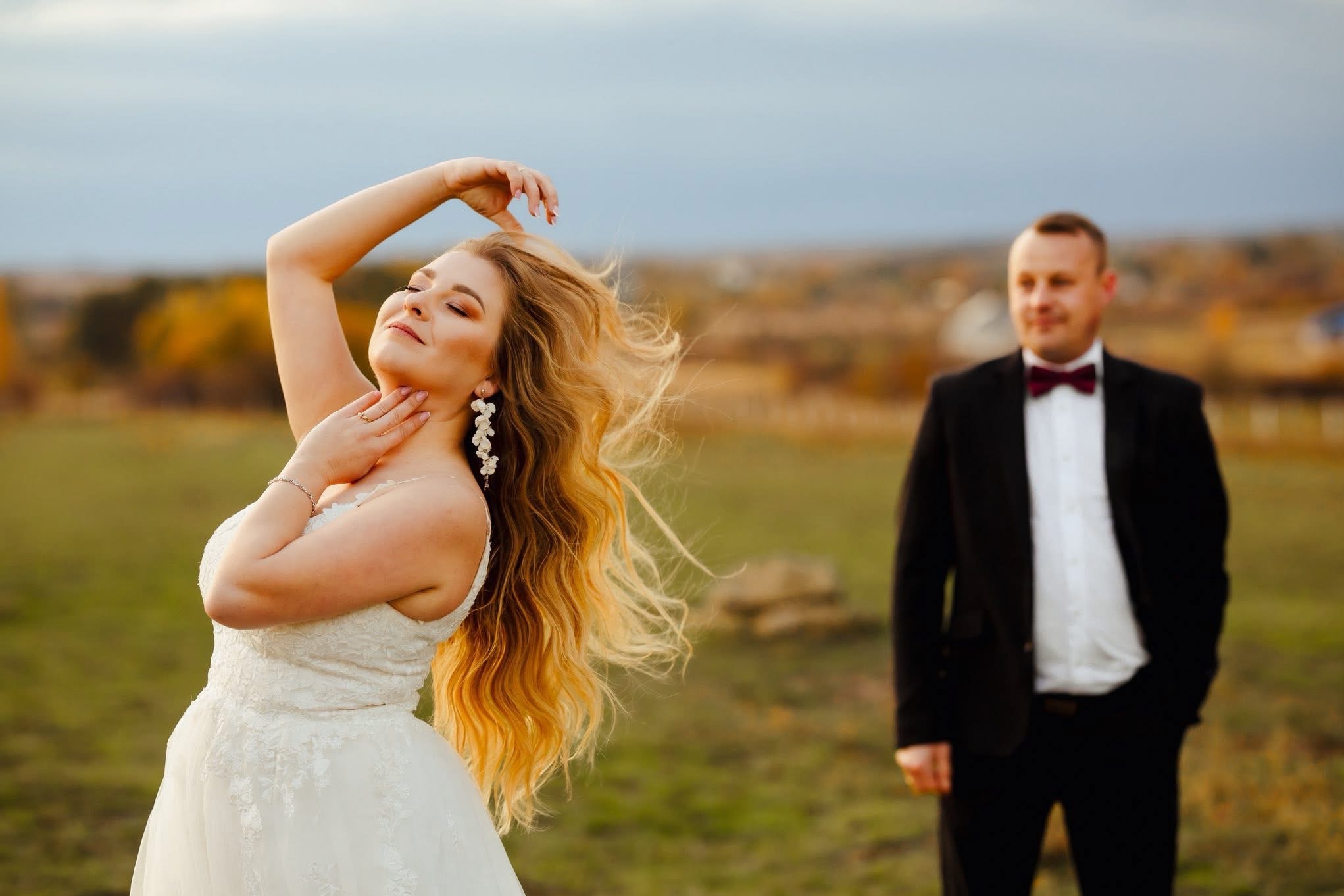 Para w plenerze: panna młoda w koronkowej sukni pozuje z uniesionymi rękami, a pan młody w czarnym garniturze z bordową muchą stoi w tle.