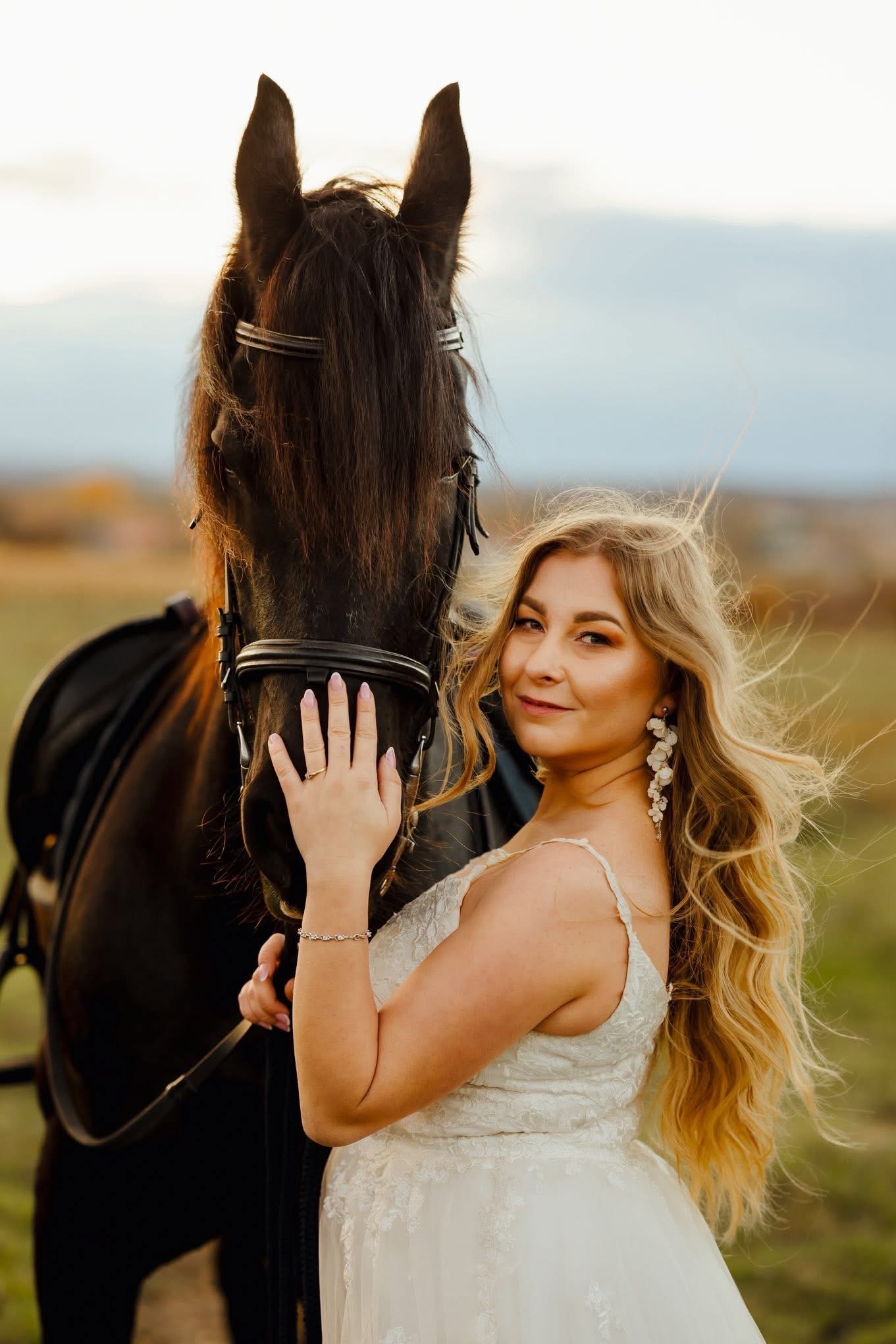 Uśmiechnięta blondynka w białej sukni pozuje z czarnym koniem na tle łąki. Delikatne światło podkreśla detale sukni i biżuterii. Wietrzna pogoda.