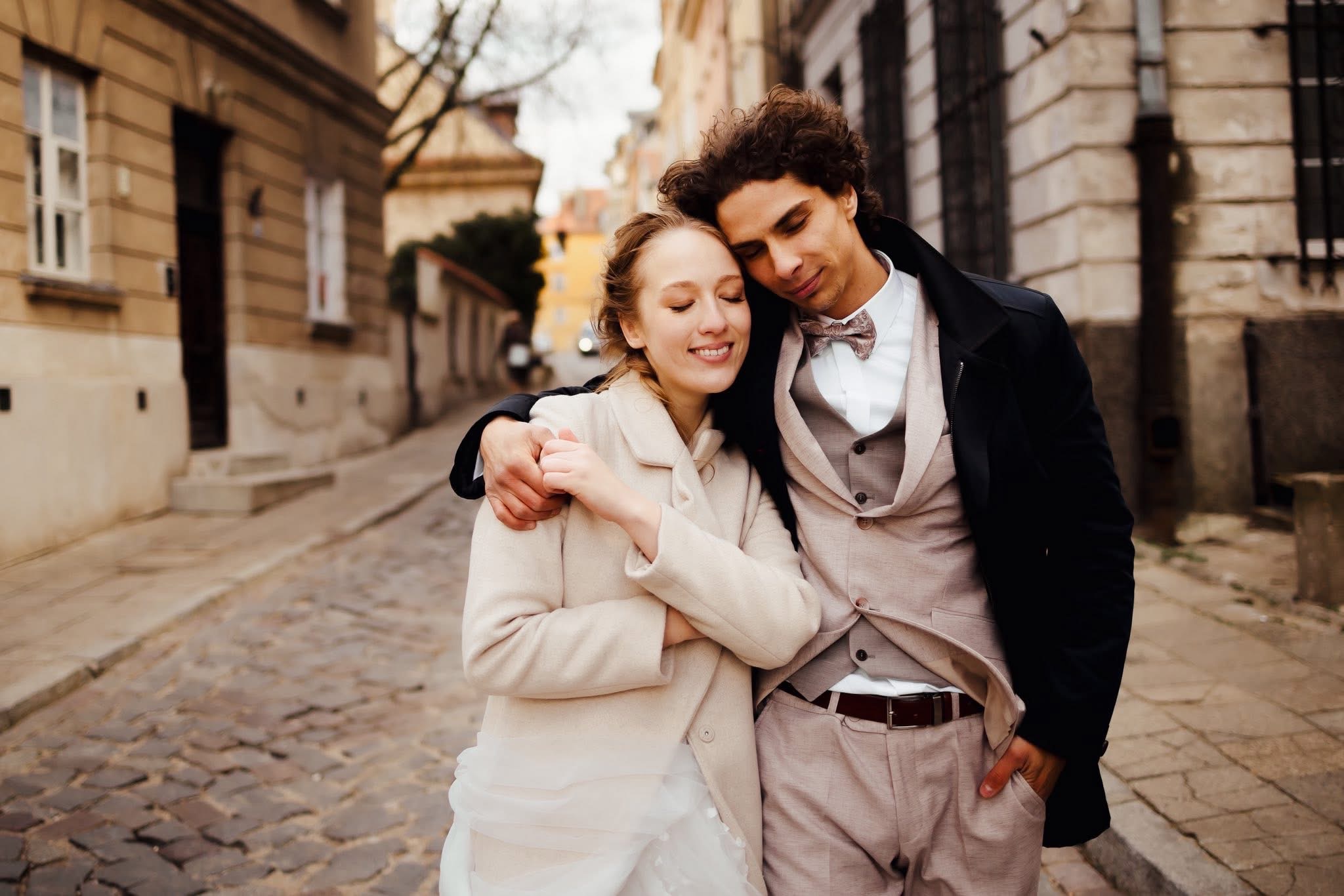 Uśmiechnięta para młoda w eleganckich strojach, obejmująca się na brukowanej uliczce w otoczeniu starych kamienic. Romantyczna sesja narzeczeńska w miejskiej scenerii.