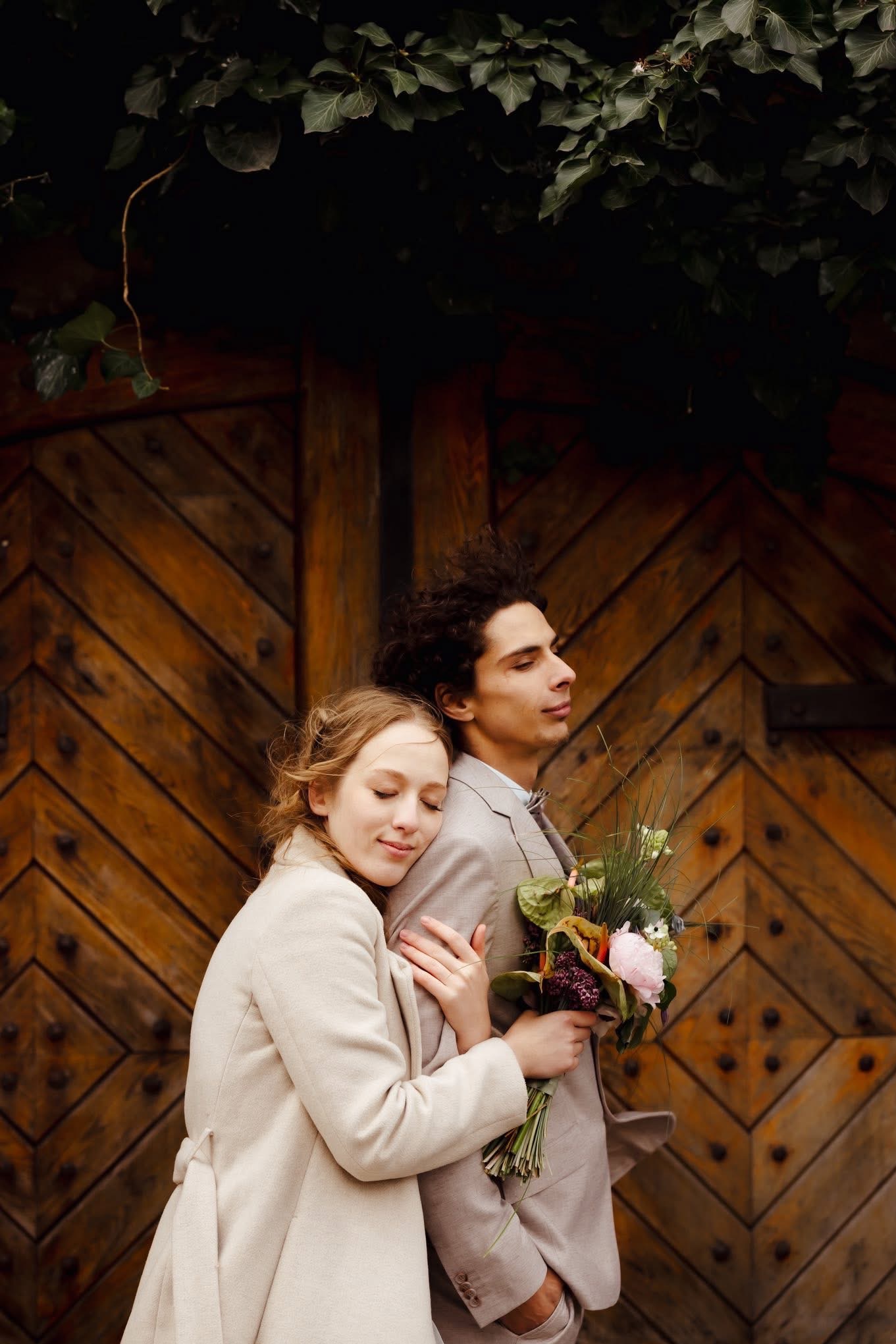 Para młoda w objęciach na tle rustykalnych, drewnianych drzwi z dekoracją roślinną. Delikatne światło podkreśla intymność chwili. Bukiet w dłoniach.
