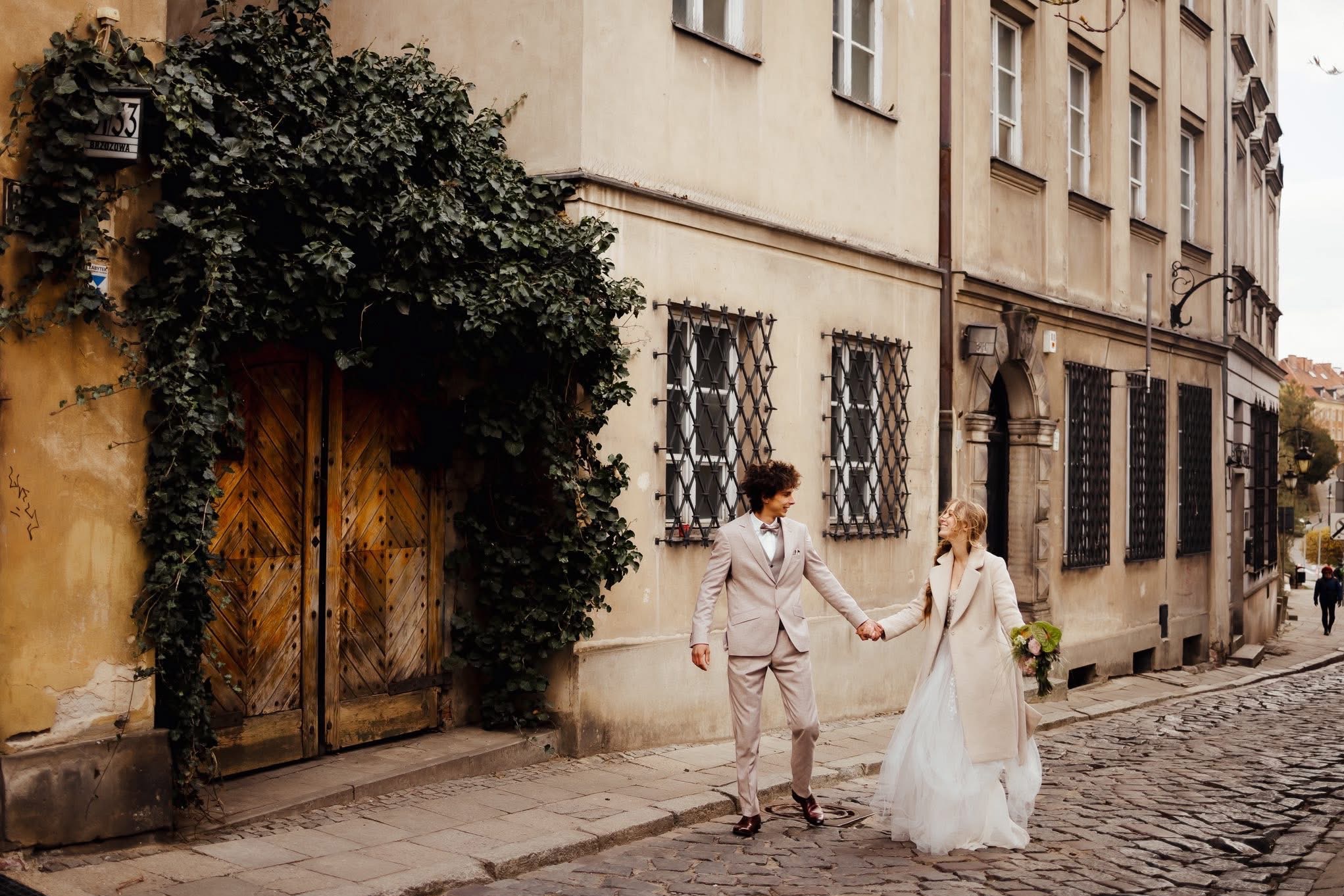 Para młoda trzymająca się za ręce na brukowanej uliczce Starego Miasta, obok kamienicy z drewnianymi drzwiami obrośniętymi bluszczem i ozdobnymi oknami.
