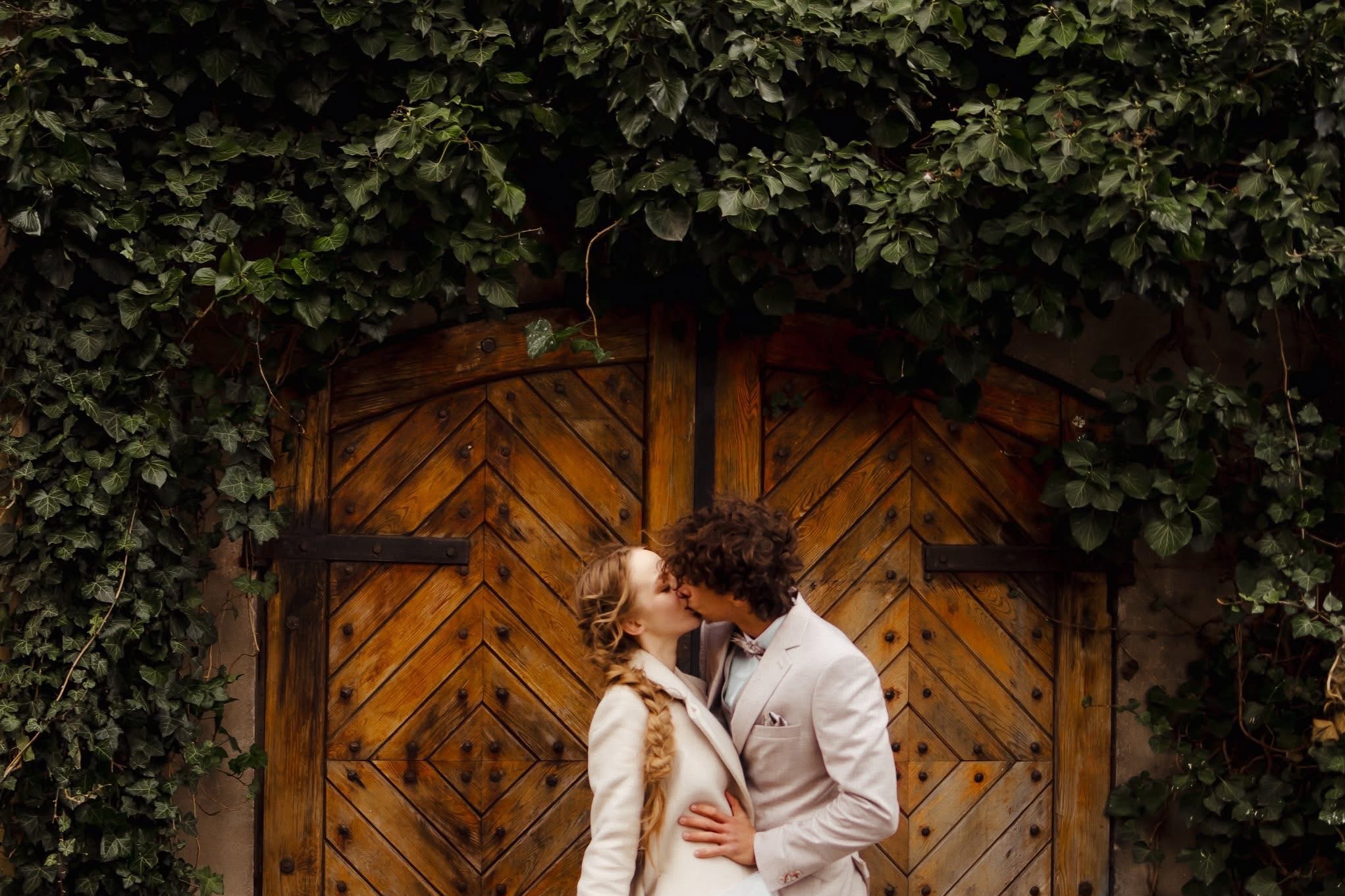 Zakochana para w pastelowych strojach całuje się na tle starych, drewnianych wrót obrośniętych bluszczem. Romantyczna sesja w plenerze, ciepłe światło.