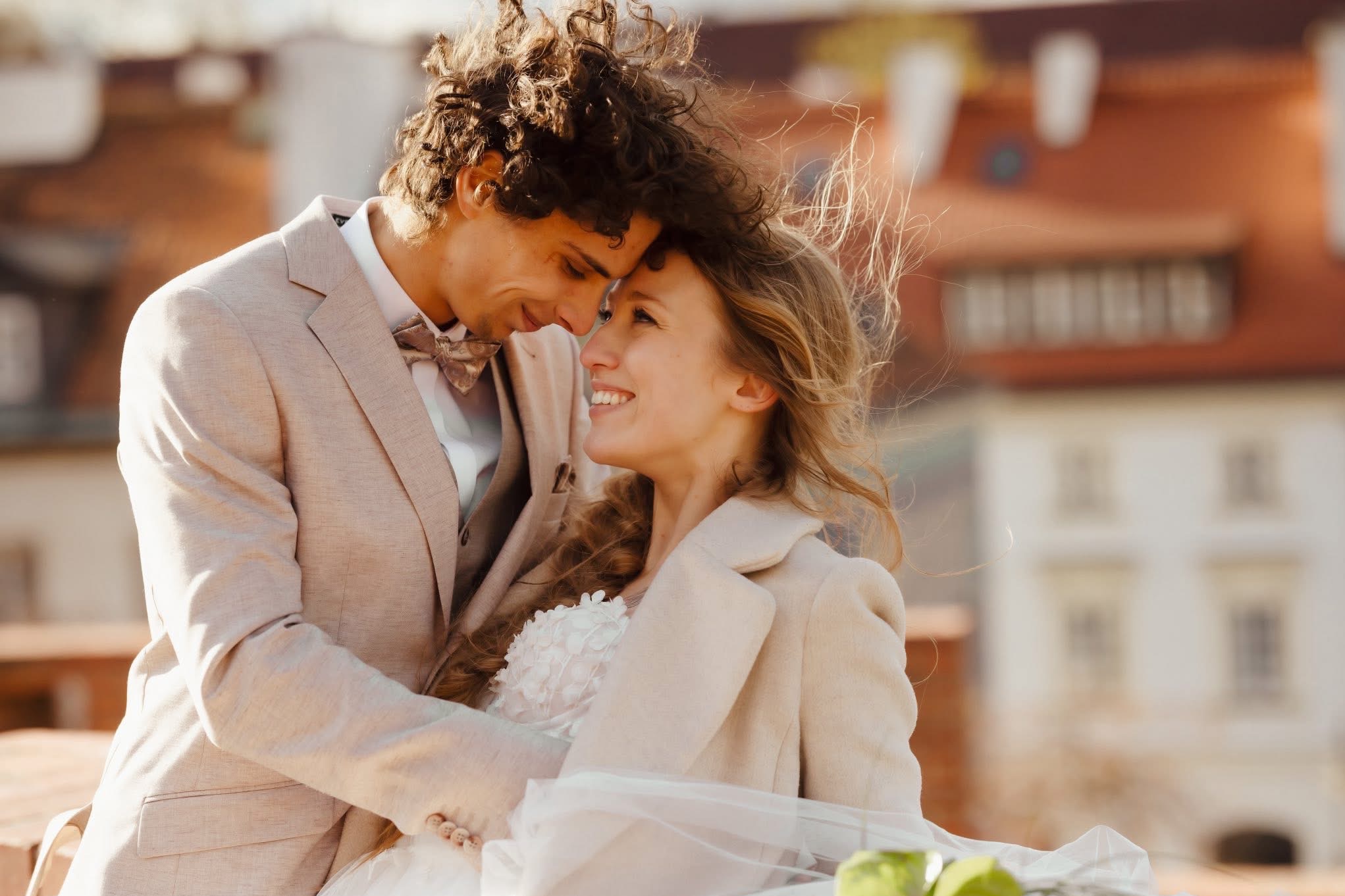 Uśmiechnięta para młoda w beżowych strojach, przytulona na tle miejskiej zabudowy z czerwonymi dachami. Delikatne światło podkreśla romantyczny nastrój.