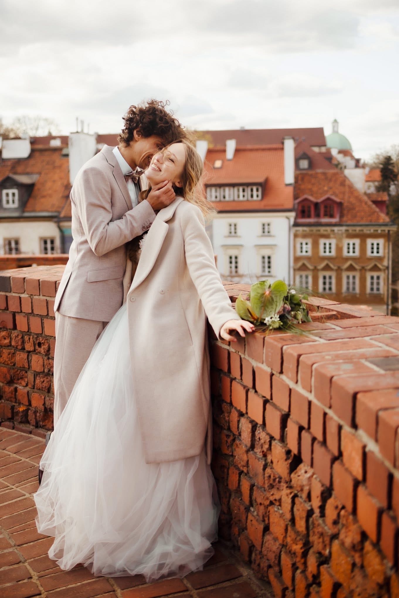Uśmiechnięta panna młoda w płaszczu, oparta o ceglany mur, z bukietem. Obok pan młody w beżowym garniturze, obejmujący ją czule. W tle zabudowa miejska.
