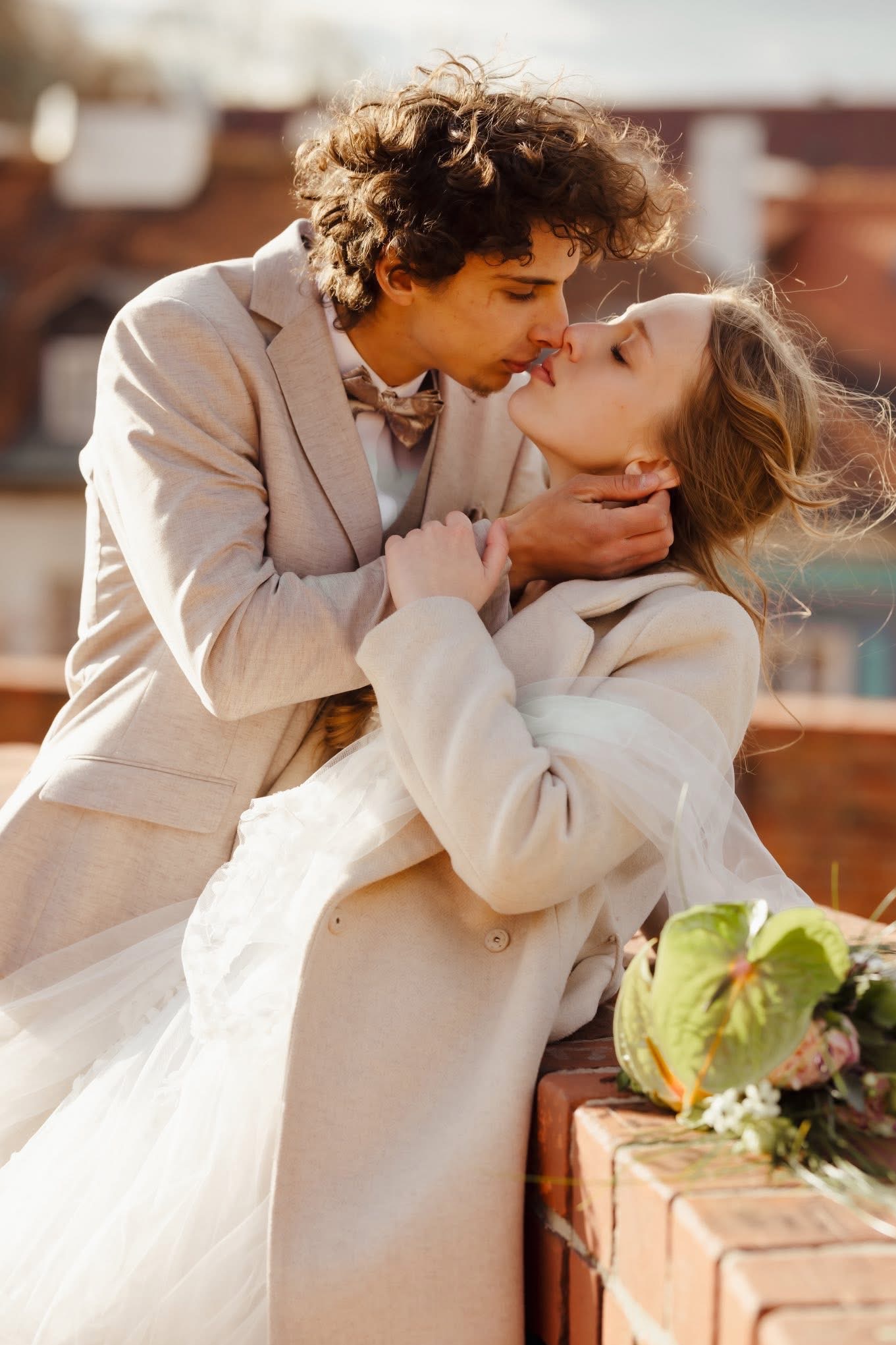 Para młoda w beżowych strojach, delikatny pocałunek na tle ceglanej balustrady i bukietu. Romantyczna sesja w plenerze, subtelne światło podkreśla intymność chwili.
