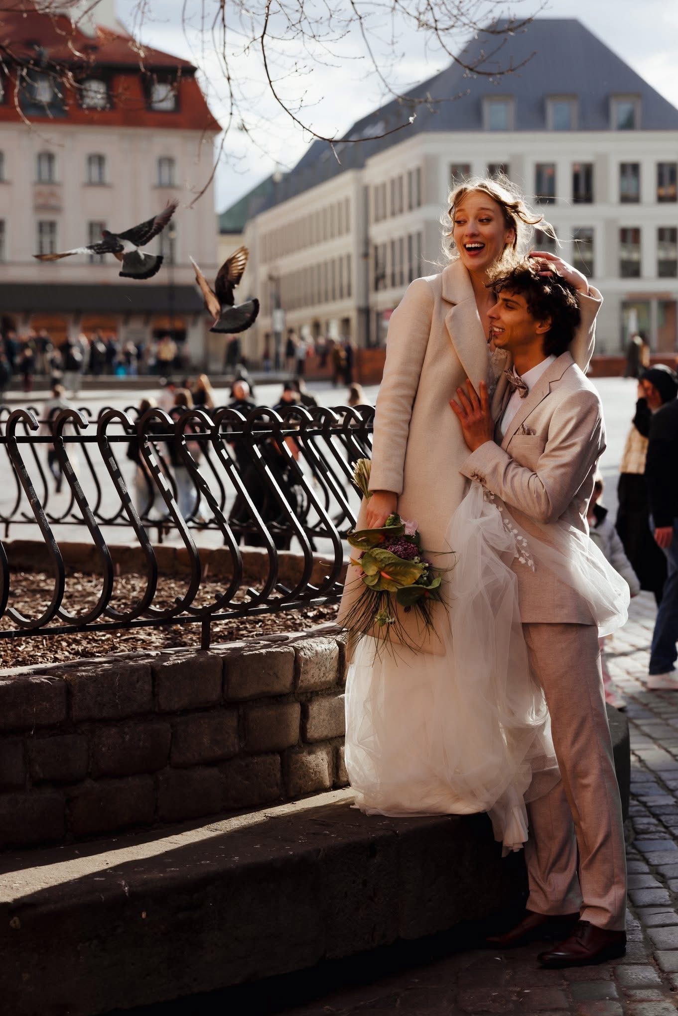 Uśmiechnięta para młoda w jasnych strojach na tle miejskiego placu z gołębiami w locie. Panna młoda trzyma bukiet. Ujęcie w słoneczny dzień.