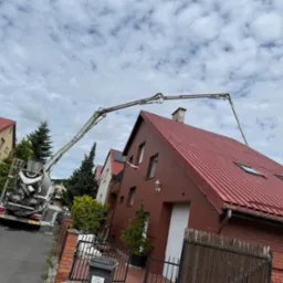 Betonomieszarka z wysięgnikiem pompuje beton na dach domu z czerwoną blachodachówką i oknami dachowymi, widok z ulicy w pochmurny dzień.