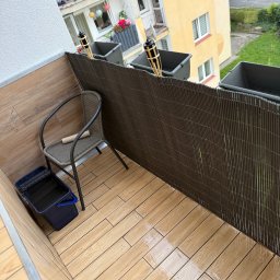 Adnel - Wykończony balkon z drewnopodobnymi płytkami, brązową osłoną i krzesłem. Widoczny kubeł z akcesoriami malarskimi. Dekoracyjne lampiony w donicach.