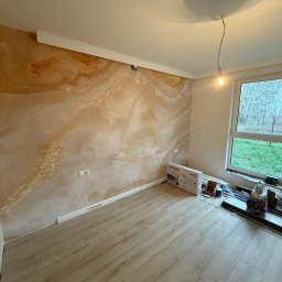 Adnel - Pokój po wykończeniu: dekoracyjna ściana imitująca marmur, jasna podłoga z paneli, białe listwy przypodłogowe i sufitowe. Widoczne narzędzia i materiały budowlane.