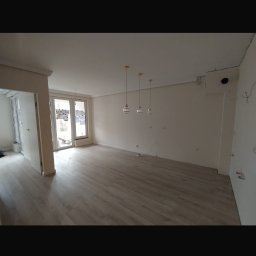 Adnel - Jasne panele podłogowe w przestronnym, odświeżonym pokoju z trzema wiszącymi lampami. Świeżo pomalowane ściany i duże okno balkonowe w tle. Widoczne ślady po instalacji elektrycznej.