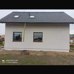Adnel - Otynkowany dom jednorodzinny z brązowymi oknami i grafitowym dachem. Widoczne dwa okna dachowe. Żółta rura spustowa. Częściowo zagospodarowany teren wokół budynku.