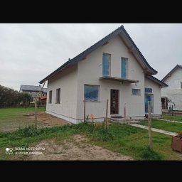 Adnel - Dom jednorodzinny po remoncie elewacji w Koszalinie. Widoczne zabezpieczone okna folią malarską. Świeżo pomalowana fasada w jasnym odcieniu.