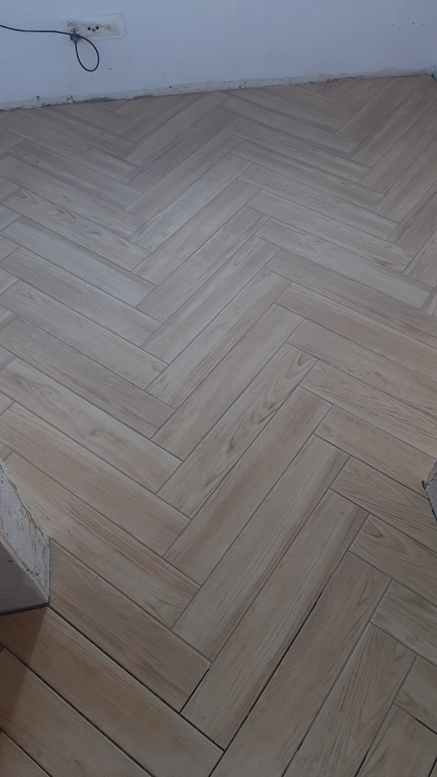 Ułożona pod kątem jasna podłoga z płytek imitujących drewno, widoczny fragment ściany z gniazdkiem elektrycznym.