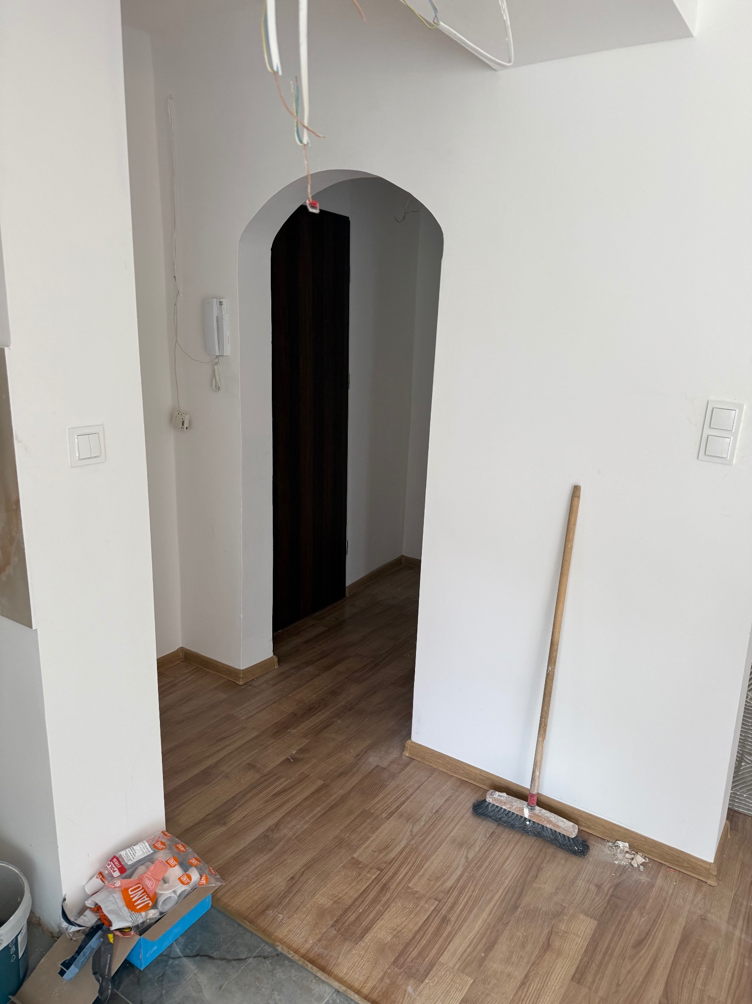 Jasne wnętrze po remoncie: świeżo pomalowane ściany, ułożone panele podłogowe. Widoczna zamiataczka i materiały budowlane, łukowate przejście do ciemniejszego pomieszczenia.