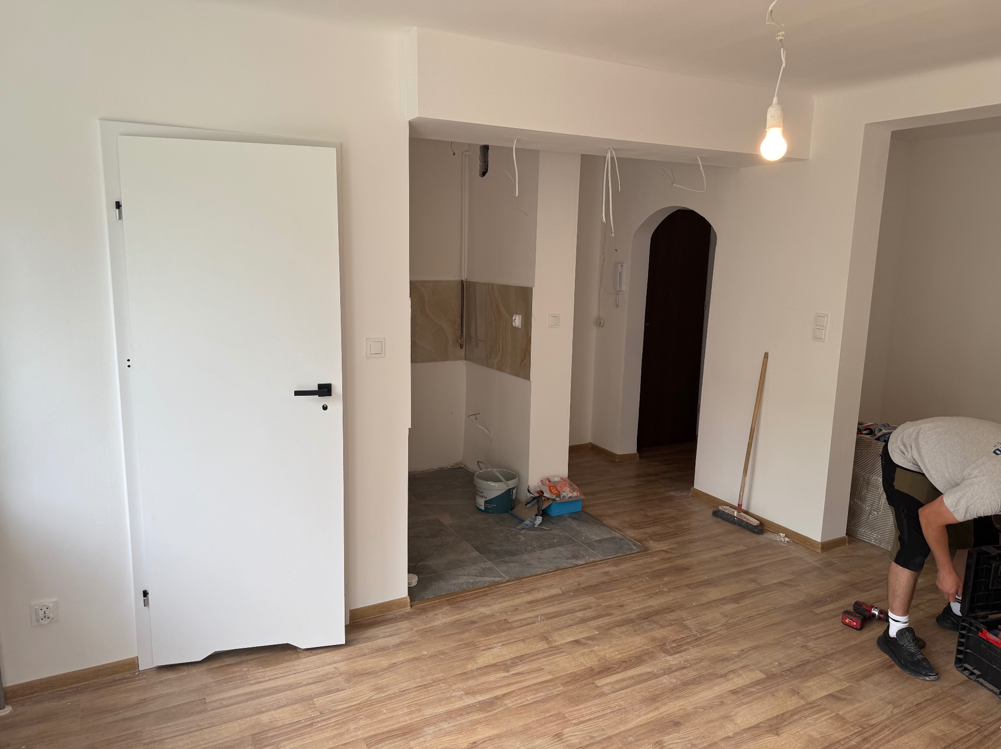 Wnętrze po remoncie: świeżo ułożone panele podłogowe, pomalowane ściany na biało, widoczne narzędzia i fragment osoby przy pracy, surowa kuchnia w tle.