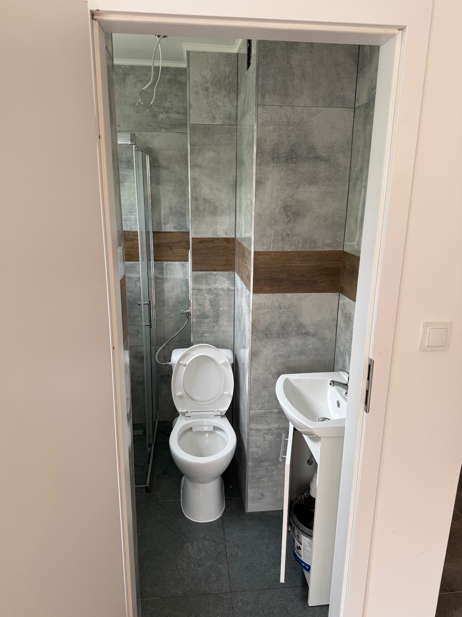 Wykończona łazienka z szarymi płytkami, drewnianym pasem dekoracyjnym i kabiną prysznicową za toaletą. Umywalka z szafką. Widok zza otwartych drzwi.